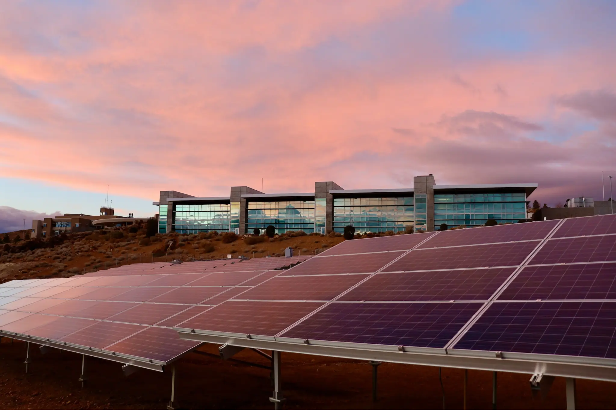 An image of solar panels under a sunset with a building behind the panels.